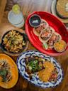 A top view of a table with various dishes: tacos with pico de gallo and rice, grilled chicken with rice, macaroni and cheese with jalapeños, a salad, cornbread, and a lime-garnished drink.