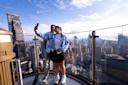A couple takes a selfie on an outdoor observation deck, surrounded by towering skyscrapers and a vast cityscape under a clear blue sky.