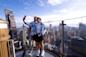 A couple takes a selfie on an outdoor observation deck, surrounded by towering skyscrapers and a vast cityscape under a clear blue sky.