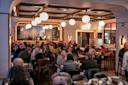 A stylish, modern restaurant filled with people dining at tables. Warm lighting from globe lamps and wall sconces creates a cozy atmosphere. A large mirror reflects the lively crowd and decor.
