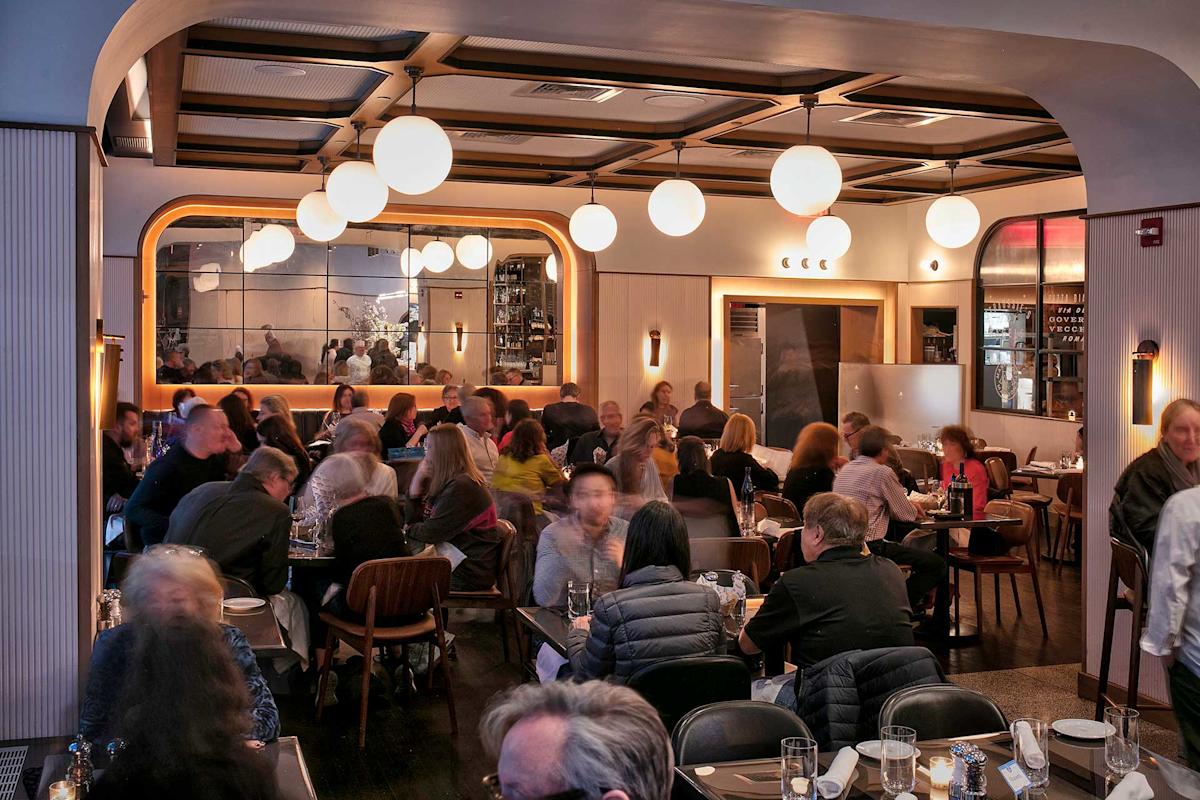 A stylish, modern restaurant filled with people dining at tables. Warm lighting from globe lamps and wall sconces creates a cozy atmosphere. A large mirror reflects the lively crowd and decor.