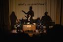 A stage performance features three people seated, with dramatic shadow projections on the backdrop. A person in the center interacts with props on a table. The words "what's on your mind?" are displayed above them.