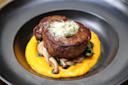 A gourmet dish featuring a juicy steak topped with herbed butter, resting on a bed of sautéed mushrooms and greens. It's served over smooth carrot puree, all presented elegantly on a black plate.