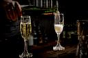 A person pours sparkling wine from a green bottle into a champagne flute next to another filled glass, on a dark wooden bar with bottles blurred in the background.