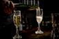 A person pours sparkling wine from a green bottle into a champagne flute next to another filled glass, on a dark wooden bar with bottles blurred in the background.
