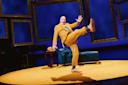 A performer in a bright yellow suit strikes a playful pose with one leg kicked high on a stage with a blue couch, yellow rug, and empty picture frames against a blue backdrop.