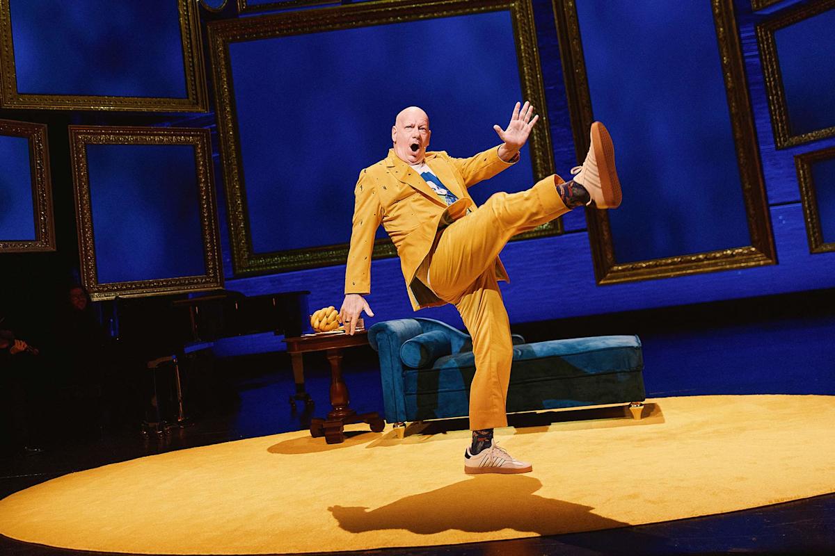 A performer in a bright yellow suit strikes a playful pose with one leg kicked high on a stage with a blue couch, yellow rug, and empty picture frames against a blue backdrop.