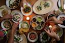 A tabletop filled with various Mexican dishes, including tacos, ceviche, and sauces. Several hands reach in to serve or sample the colorful food. Candles provide a warm ambiance, and drinks are visible at the edges of the table.