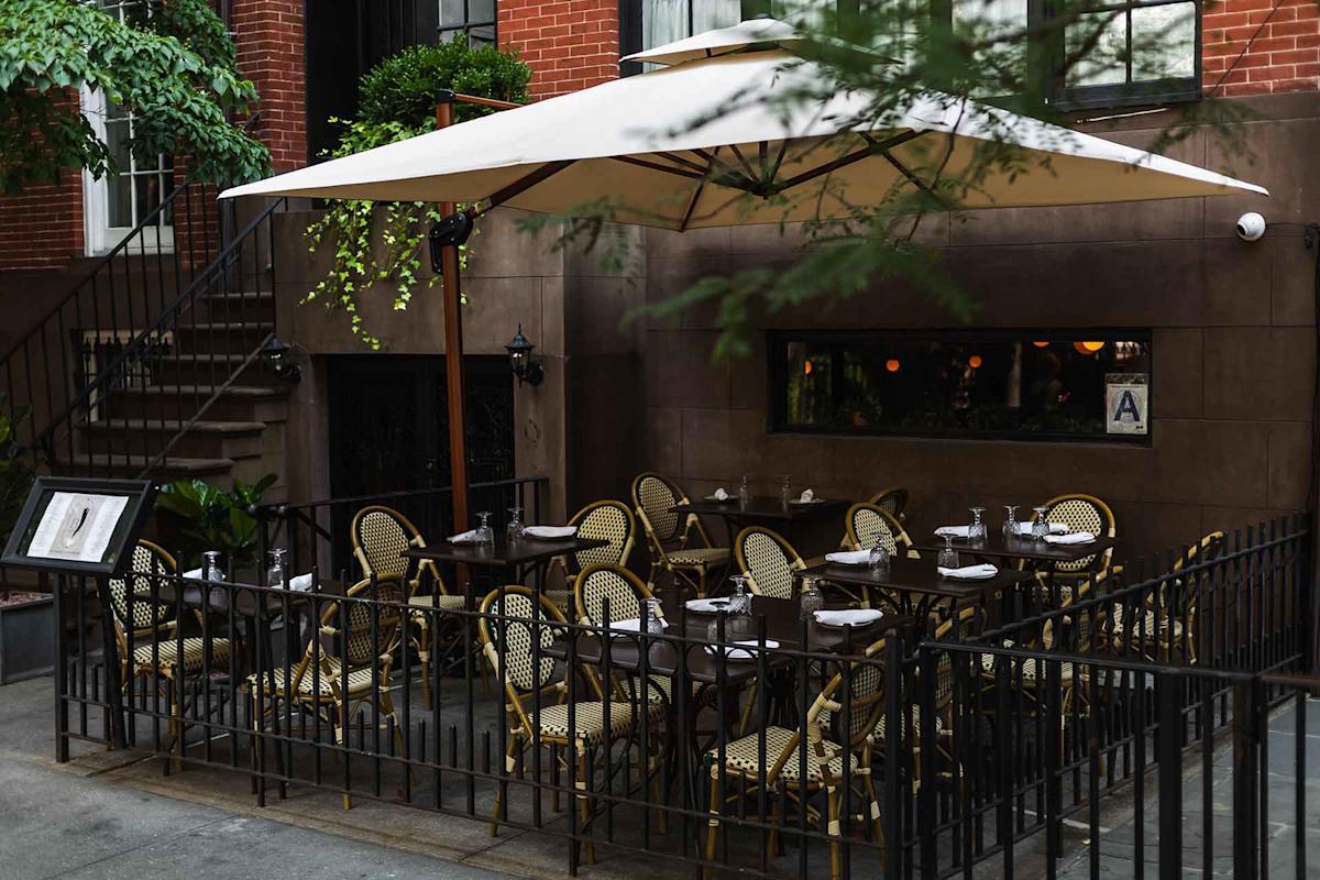 Outdoor patio of a restaurant with empty woven chairs and tables, surrounded by a black metal fence, under a large white umbrella, next to a brick building.