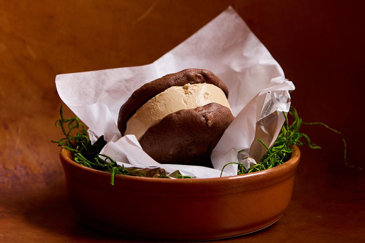 A chocolate ice cream sandwich with vanilla filling sits on white parchment paper in a brown dish, surrounded by decorative green moss against a brown background.