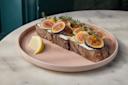 A slice of rustic bread topped with fresh figs, creamy spread, honey, and herbs sits on a pink plate with a lemon wedge on the side, placed on a marble table.