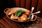 A baked or roasted chicken cut into pieces is served in an oval dish, garnished with fresh greens, with serving utensils placed inside. The dish sits on a wooden table.