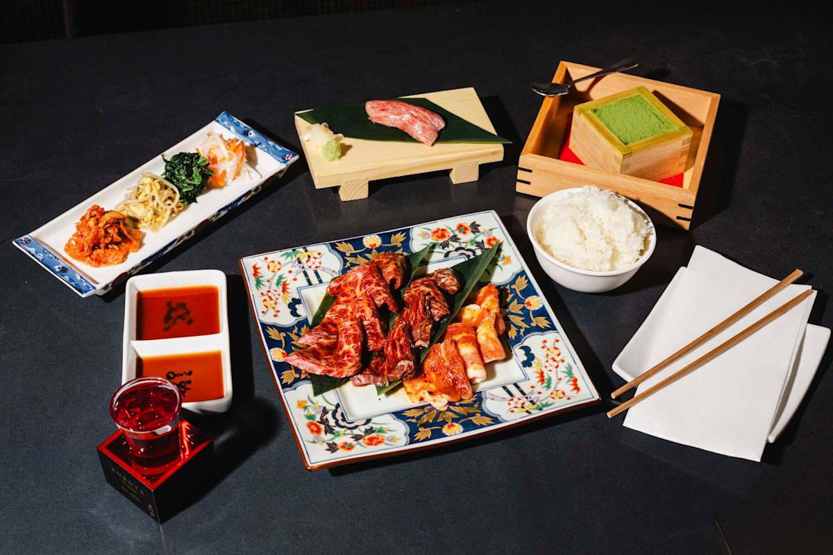An assortment of Japanese dishes on a table, including a plate of grilled meat, a bowl of rice, pickled vegetables, green tea dessert, and a small cup with a red liquid. Chopsticks and a napkin are placed nearby.