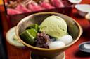 A bowl with green tea ice cream, green jelly cubes, red bean paste, and mochi balls. The background shows a dish of sliced meat, slightly out of focus.