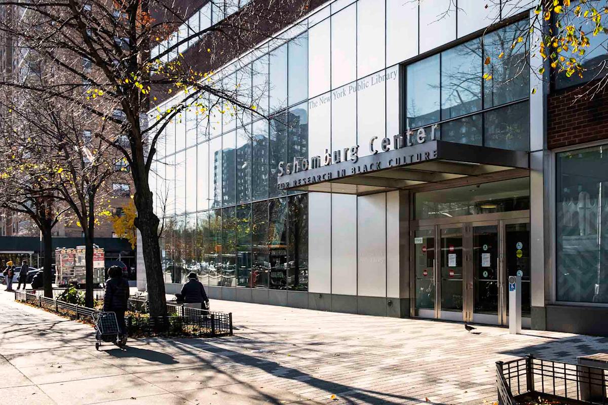 Street view of the Schomburg Center for Research in Black Culture, featuring a glass façade with the name displayed at the entrance. Pedestrians and leafless trees line the sidewalk under a clear sky.