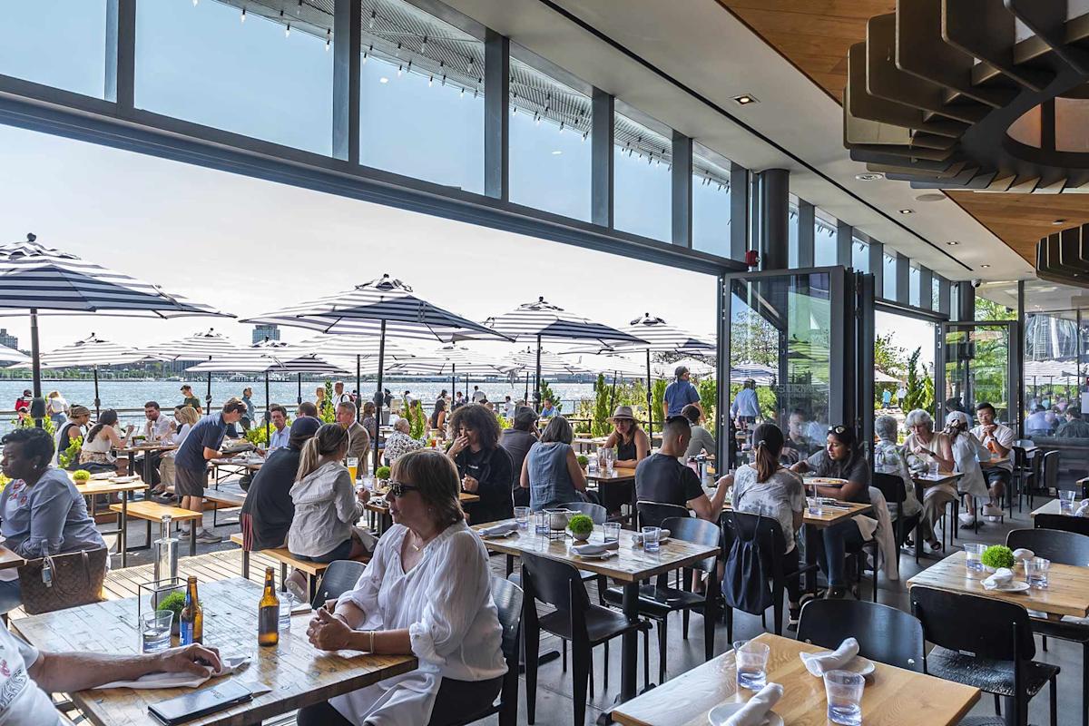 People dine at a busy, modern restaurant with large windows and outdoor seating. Black-and-white striped umbrellas shade the patio; sunlight streams in, and greenery surrounds the waterfront view.