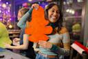 A smiling person holds a large orange shape resembling a paper cutout in a lively indoor setting. Another person beside them is focused on crafting tasks. Decor and colorful lights are visible in the background.