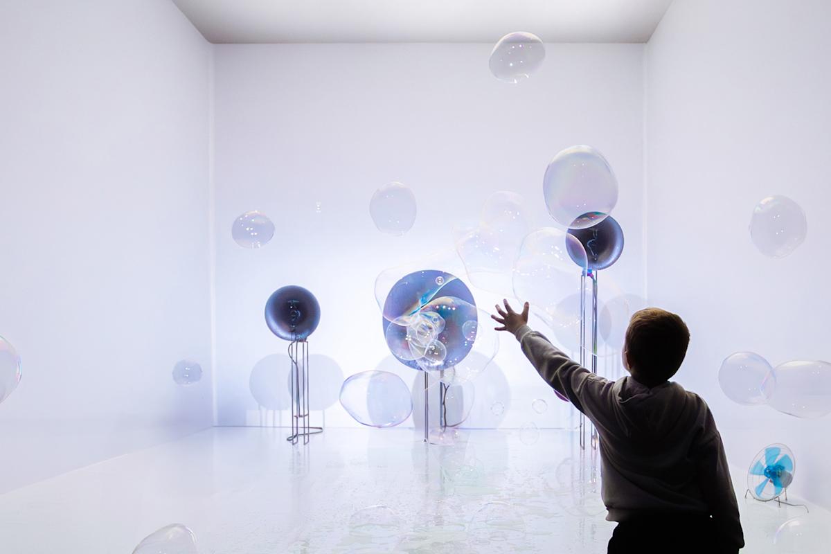 A child reaches out to touch floating bubbles in a bright, white room filled with art installations made of large transparent spheres on stands.