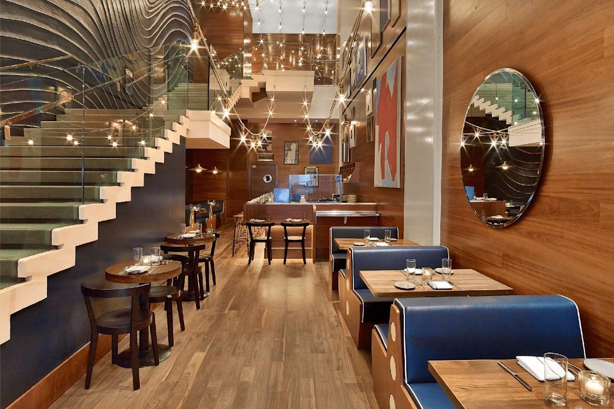 A modern restaurant interior with wooden floors, dark wood tables, and blue booth seating. String lights hang from the ceiling, and a staircase with a glass railing leads to an upper level. Round mirrors and art decorate the walls.