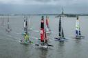Several colorful sailboats race on a gray, cloudy day in a wide harbor, with the Statue of Liberty visible in the background on the right side.