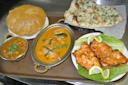 A tray with various Indian dishes: a puffed puri, a bowl of chickpea curry, a dish of creamy curry with herbs, two pieces of grilled chicken with lemon on lettuce, and a basket of garlic naan.