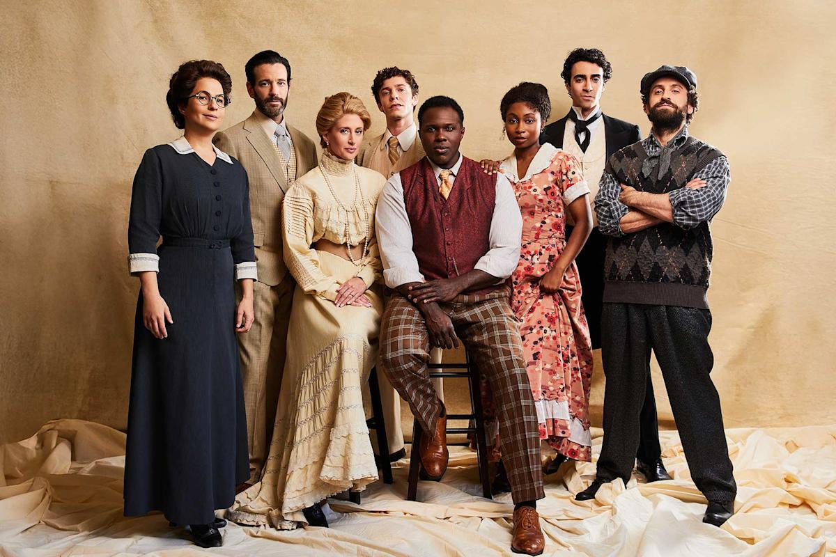 Eight people in period clothing pose together against a beige backdrop; one man sits center while others stand around him, wearing vintage dresses, suits, and vests, evoking an early 20th-century style.