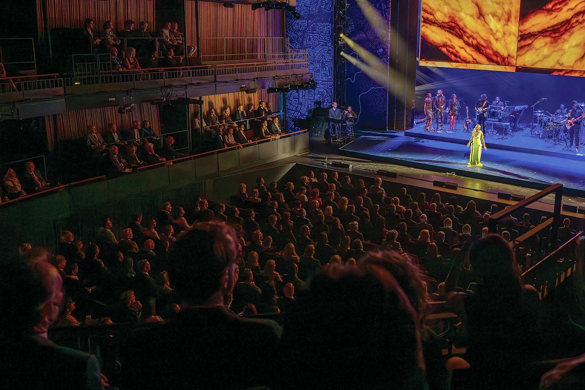A singer in a yellow dress performs on a brightly lit stage with musicians and backup singers, while a large audience watches from a darkened theater.