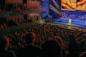 A singer in a yellow dress performs on a brightly lit stage with musicians and backup singers, while a large audience watches from a darkened theater.