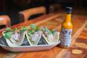 Three fish tacos topped with cilantro and sliced radishes are served on a rectangular white plate next to a bottle of Palo Santo salsa picante on a wooden table.