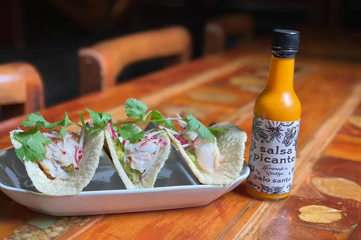 Three fish tacos topped with cilantro and sliced radishes are served on a rectangular white plate next to a bottle of Palo Santo salsa picante on a wooden table.