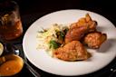 Plate of crispy fried chicken with a side of fresh vegetable slaw, served on a white dish. A glass with an orange beverage and utensils are nearby on a dark surface.