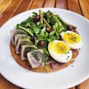 A white plate with sliced seared tuna, mixed greens, green beans, black olives, and a halved soft-boiled egg, all served on a round, golden flatbread on a wooden table.