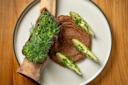 A dish featuring a bone with greens on top, served with a brown puree and sliced avocado garnished with herbs, arranged on a round white plate on a wooden table.