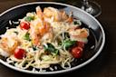 A black plate with creamy pasta topped with grilled shrimp, cherry tomatoes, spinach, and grated cheese. A glass of white wine is partly visible in the background on a dark wooden table.
