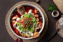 A bowl of spicy fish stew with chili oil, topped with sliced fish, fresh red and green chilies, cilantro, and Sichuan peppercorns, placed on a woven placemat with chopsticks and teaware nearby.