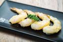 Four raw shrimp are neatly arranged on a black rectangular plate, garnished with a small sprig of parsley. The plate rests on a light wooden surface.