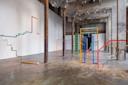 A spacious industrial gallery features colorful, minimalist stair and doorway sculptures made of metal bars, scattered across a polished concrete floor with tall columns and exposed ceilings.