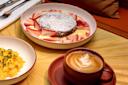 A bowl with a round chocolate cake topped with powdered sugar, surrounded by sliced strawberries and custard. Nearby are scrambled eggs with chives and a cup of coffee with latte art.