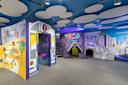 Colorful indoor children's play area featuring a bus-themed entrance and a purple tunnel structure, with playful wall murals and circular white panels on the ceiling.