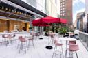 Rooftop patio with high tables and chairs, featuring a large red umbrella. A modern bar area with a wooden facade is in the background. Surrounded by tall buildings, the space is bright and inviting, filled with potted plants and city views.