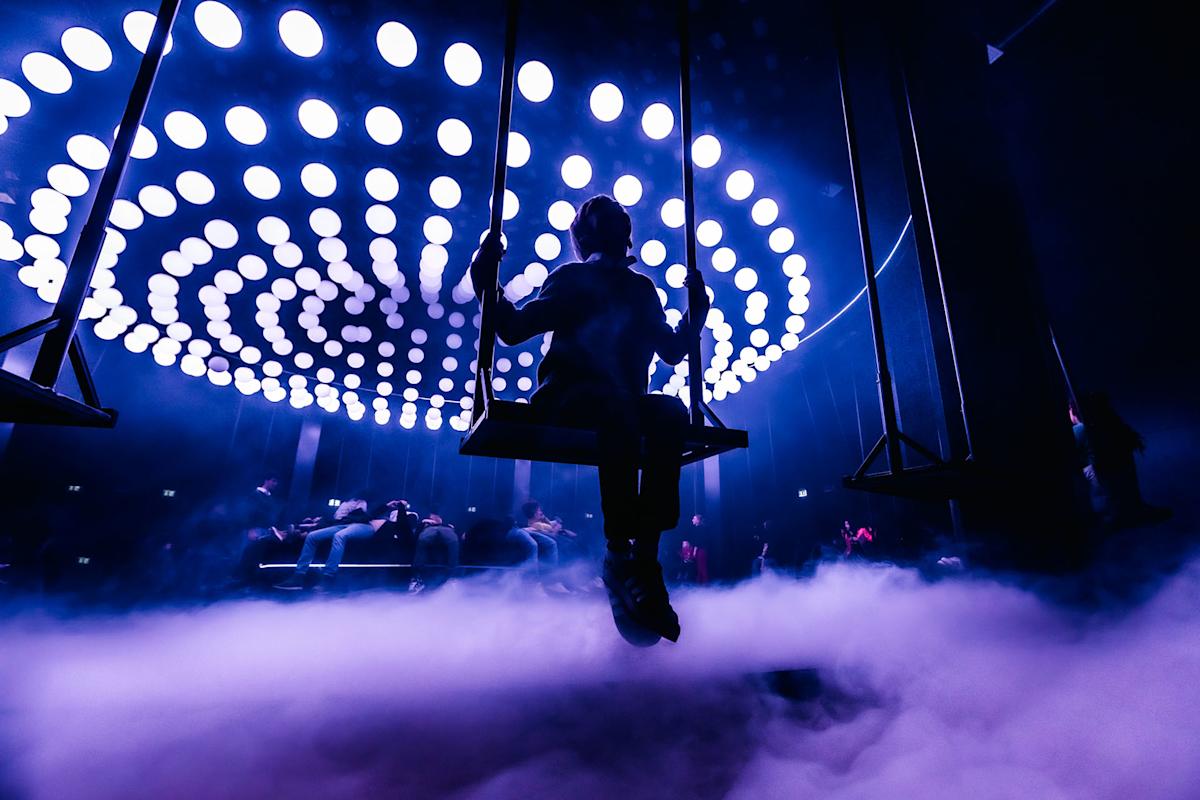 A person on a swing is silhouetted against a background of circular lights and fog, creating a dramatic and futuristic atmosphere in a dark room.