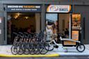 A row of rental bikes and a cargo bike are parked outside Holland Bikes, a bike rental and tour shop in NYC. The storefront features large signs, and a woman is pictured riding a bike on a poster by the entrance.