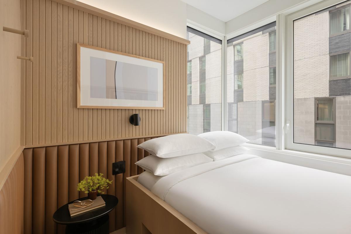 A modern hotel room with a white bed, light wood walls, a framed picture, a small black table with a plant, and large windows showing nearby buildings.