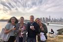 Four people smile while holding pastries by the waterfront, with a city skyline and tall buildings in the background. The sky is partly cloudy and a person with a bike is visible nearby.