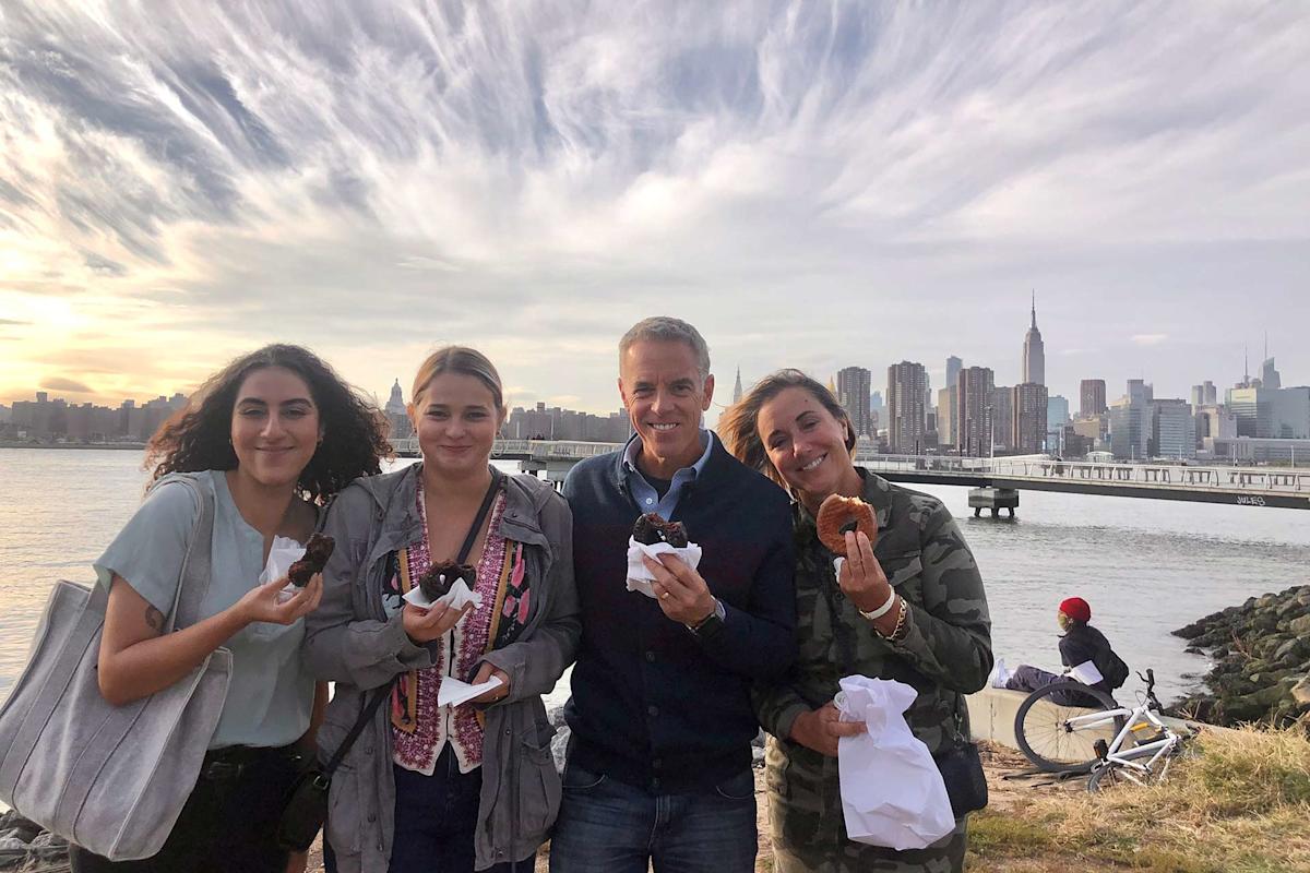 Four people smile while holding pastries by the waterfront, with a city skyline and tall buildings in the background. The sky is partly cloudy and a person with a bike is visible nearby.