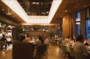A warmly lit, modern restaurant with people dining at tables, wooden floors, tall windows, and a stylish ceiling light fixture create a cozy, lively atmosphere.