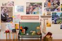 A cozy room features a green bench with a rooster statue, plants, and a tea set. The wall displays assorted photos of gardening and community activities. A "Locally Grown" sign is prominently hung among the images.