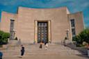The image shows the main entrance of the Brooklyn Public Library. The grand building has a large, ornate door flanked by gold detailing and two women ascending the stairs. There are green bushes and lanterns on either side of the steps.
