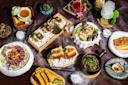 An assortment of gourmet dishes on a table includes sushi, a salad, a steak, corn on the cob, sliders, and various small side dishes. The presentation features artistic plating with decorative elements and dry ice for a smoky effect.