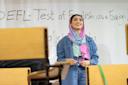 A woman wearing a colorful headscarf and denim jacket stands smiling in front of a whiteboard that reads "TEFL - Test of English as a Foreign [Language]." Classroom chairs are visible in the foreground.